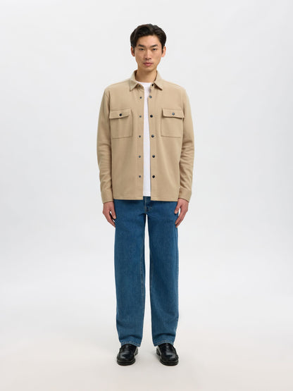 A man stands against a light gray studio background wearing a beige button-up overshirt over a white T-shirt, blue jeans, and black shoes.