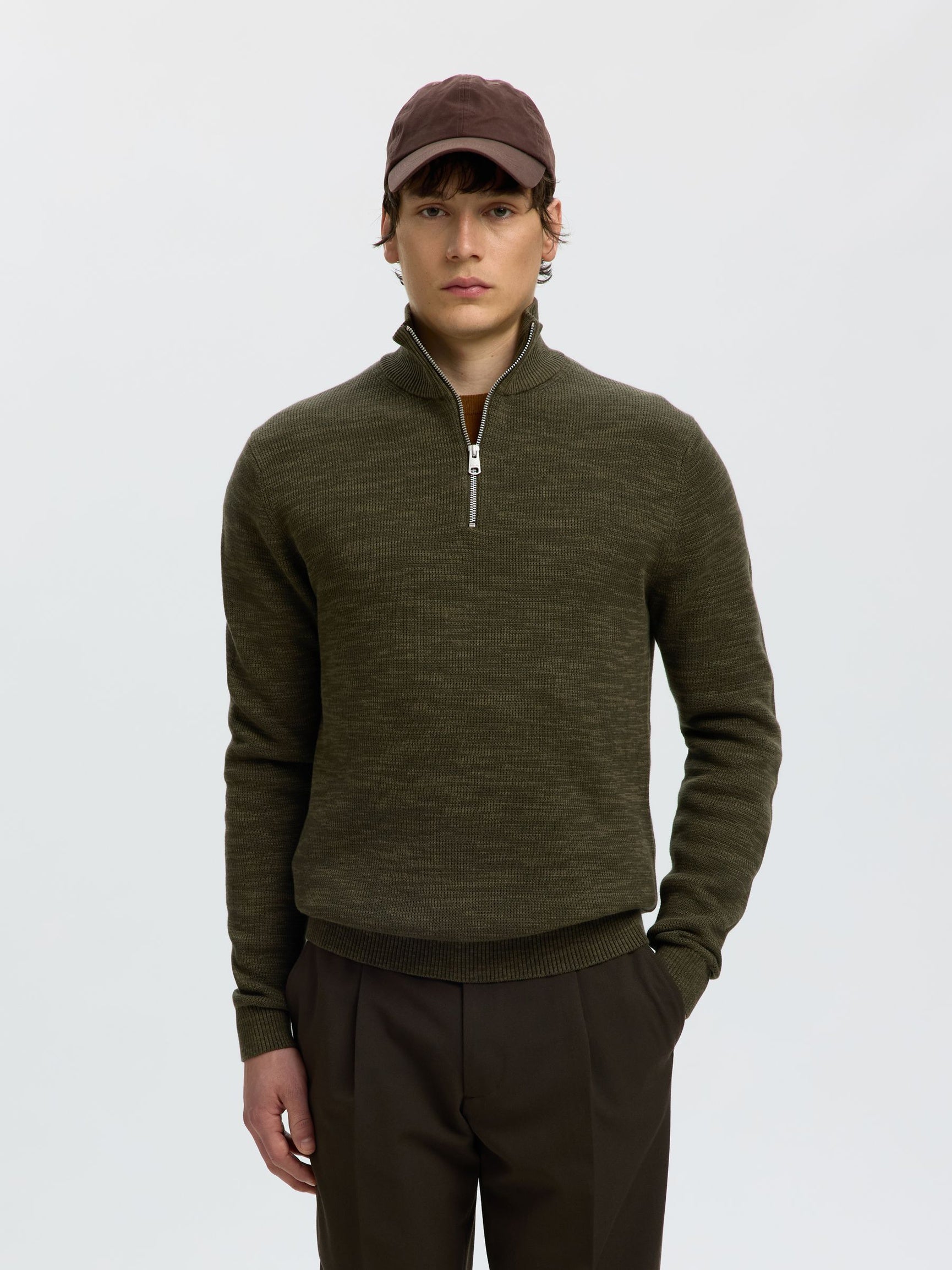 A man wearing an olive green quarter-zip knit sweater, dark brown trousers, and a dark brown baseball cap, standing against a light gray studio background.