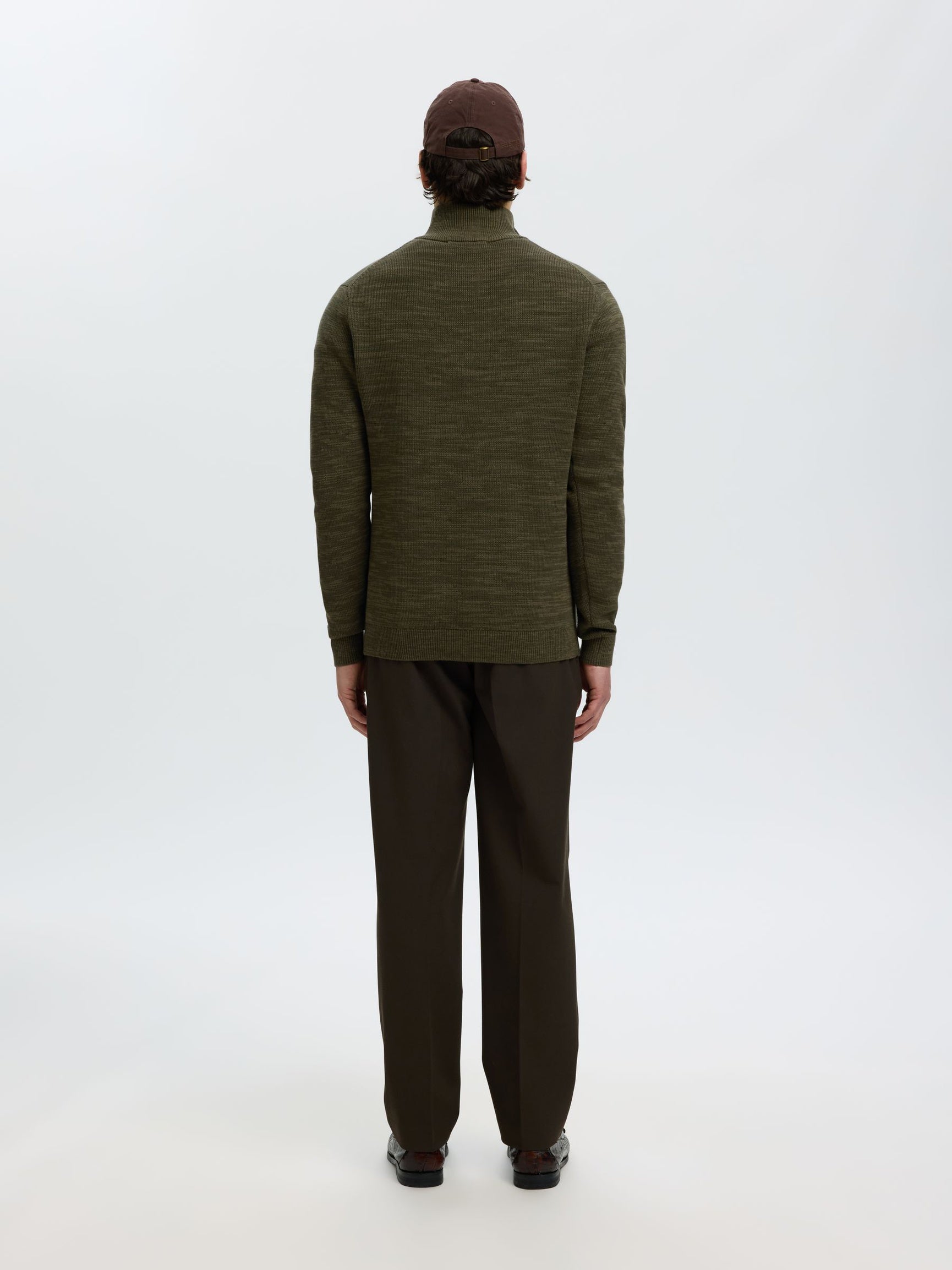 A man viewed from behind wearing an olive green long-sleeve mock neck sweater, dark brown trousers, and a dark brown cap, standing against a plain white studio background.