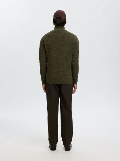 A man viewed from behind wearing an olive green long-sleeve mock neck sweater, dark brown trousers, and a dark brown cap, standing against a plain white studio background.