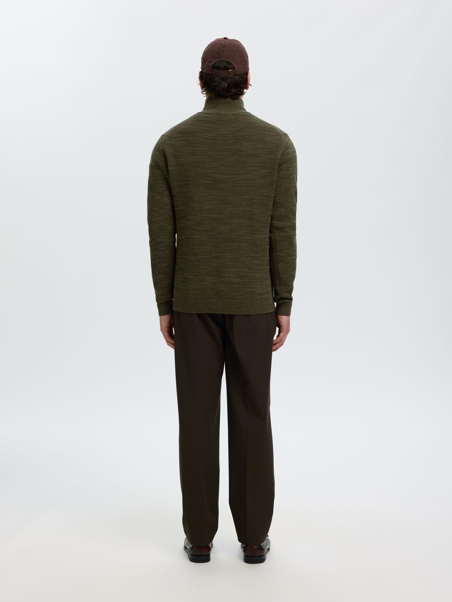 A man viewed from behind wearing an olive green long-sleeve mock neck sweater, dark brown trousers, and a dark brown cap, standing against a plain white studio background.