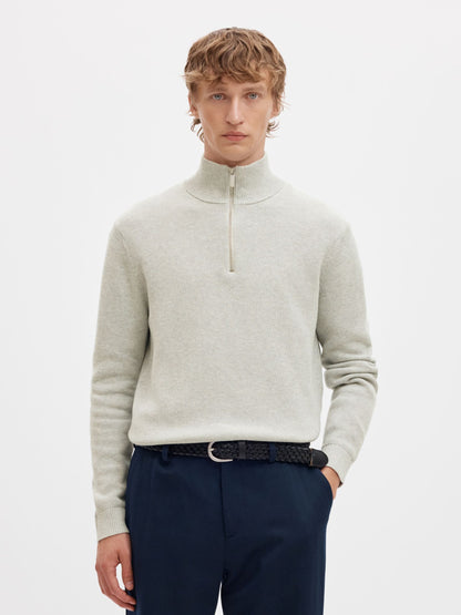 A man wearing a light beige quarter-zip mock neck sweater, navy trousers, and a black patterned belt, photographed against a clean white studio background.