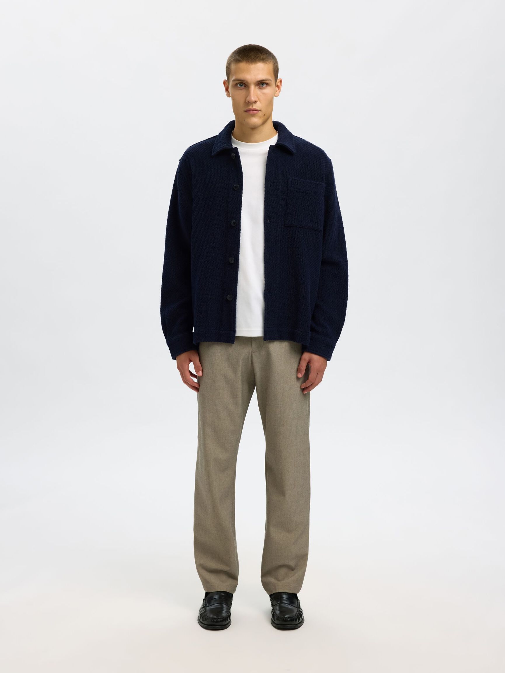 Man wearing a navy jacket, white shirt, and beige pants on a white background