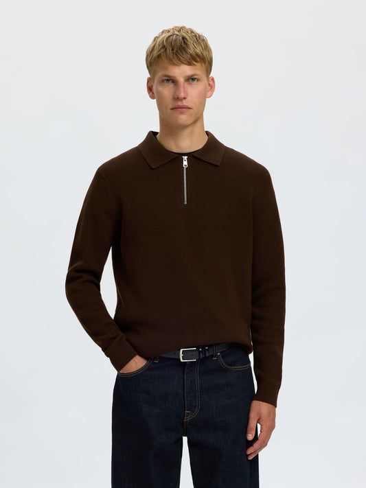A man wearing a dark brown long-sleeve polo-style knit sweater with a short front zipper, paired with dark blue jeans, against a plain light gray studio background.