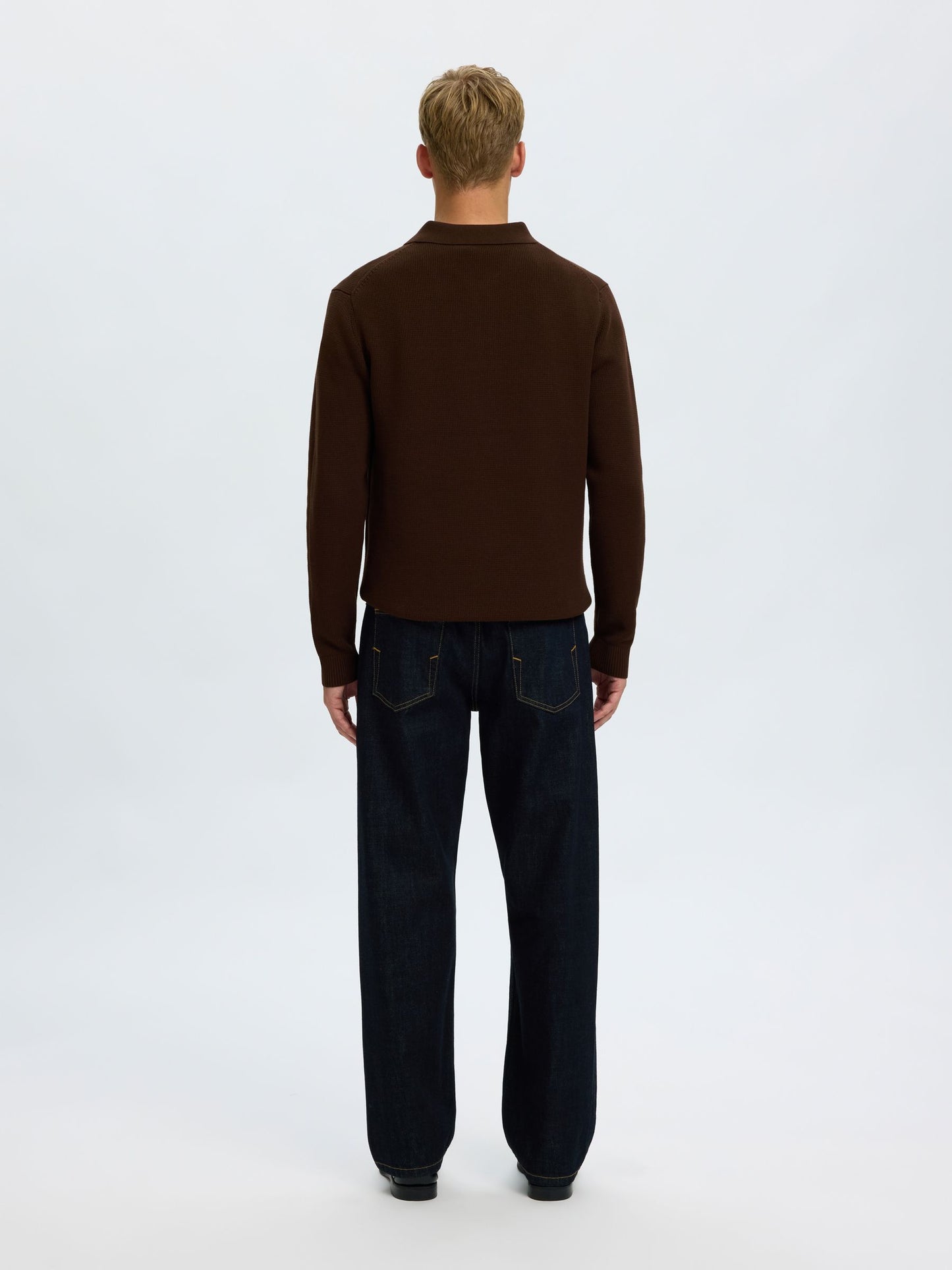 A man viewed from behind wearing a dark brown mock neck sweater and dark blue jeans, standing against a plain white studio background.