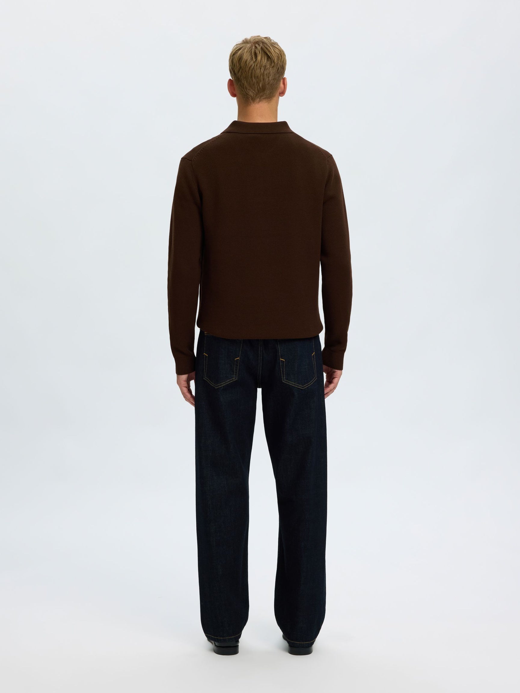 A man viewed from behind wearing a dark brown mock neck sweater and dark blue jeans, standing against a plain white studio background.