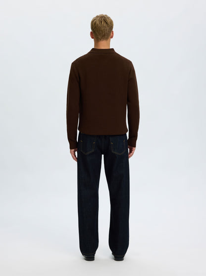 A man viewed from behind wearing a dark brown mock neck sweater and dark blue jeans, standing against a plain white studio background.