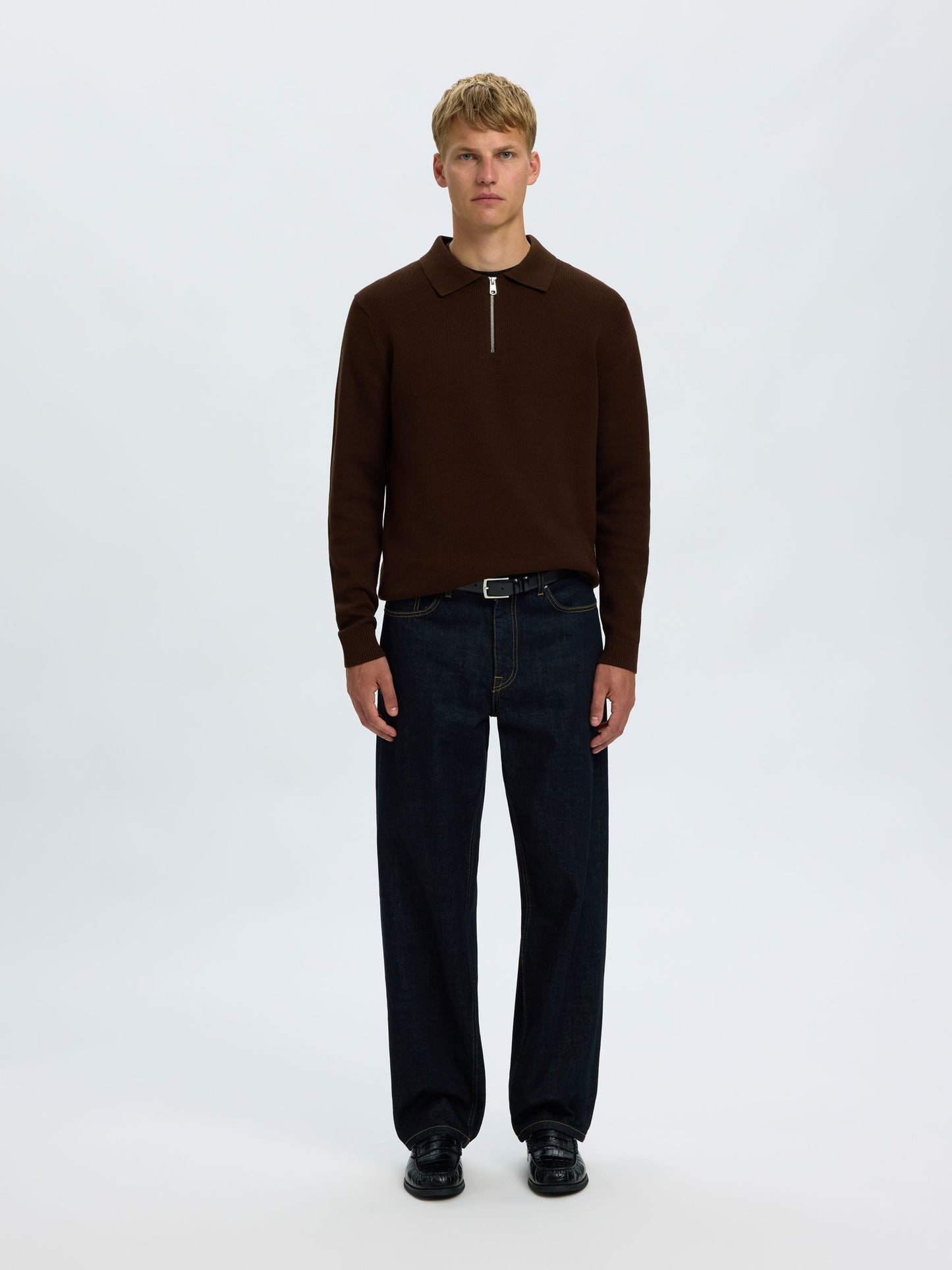 A male model stands against a light gray studio background wearing a dark brown long-sleeve knit polo-style top with a short front zipper, dark navy jeans, a black belt, and black shoes.
