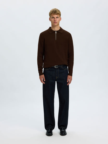 A male model stands against a light gray studio background wearing a dark brown long-sleeve knit polo-style top with a short front zipper, dark navy jeans, a black belt, and black shoes.