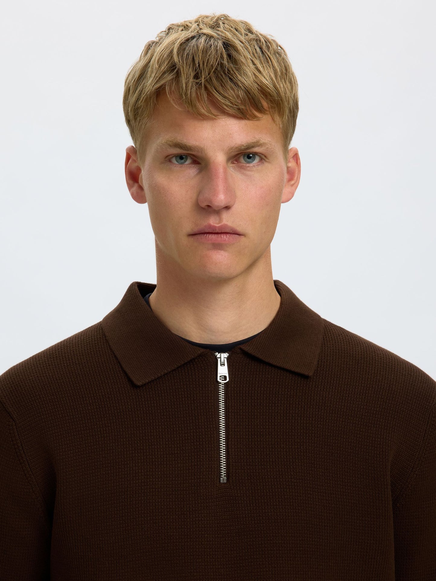 A close-up portrait of a young man with blonde hair wearing a dark brown ribbed knit polo shirt with a half-zip collar, set against a plain light gray background.