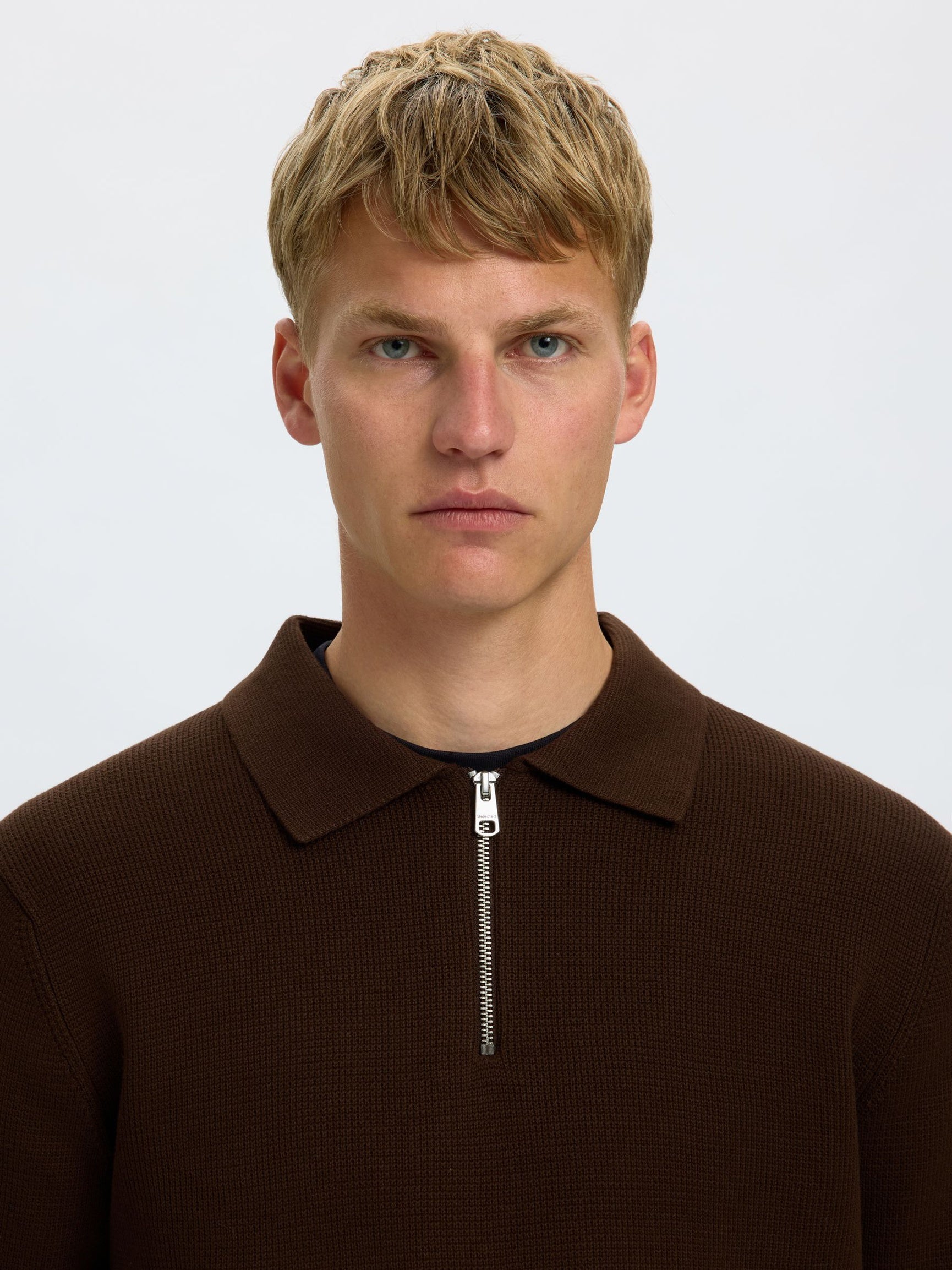 A close-up portrait of a young man with blonde hair wearing a dark brown ribbed knit polo shirt with a half-zip collar, set against a plain light gray background.