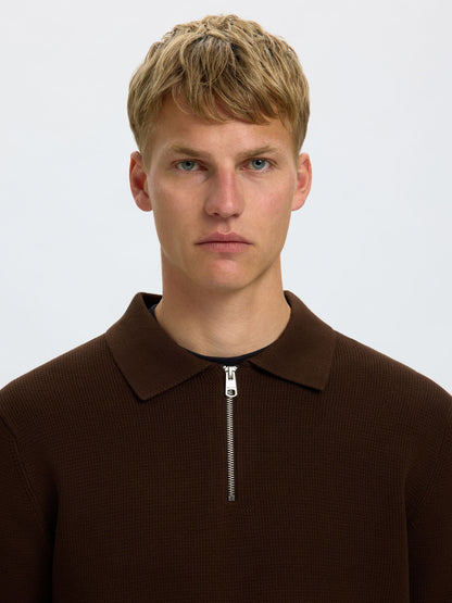 A close-up portrait of a young man with blonde hair wearing a dark brown ribbed knit polo shirt with a half-zip collar, set against a plain light gray background.