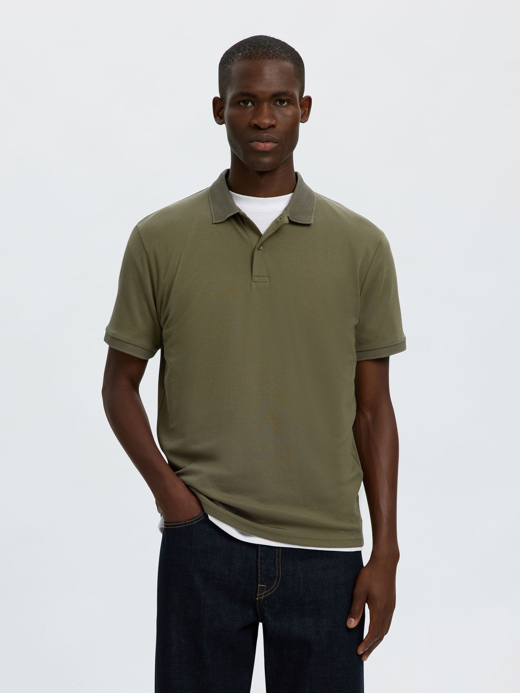 A man wearing an olive green short-sleeve polo shirt with a contrasting dark gray collar, layered over a white undershirt and paired with dark blue jeans, against a light gray studio background.