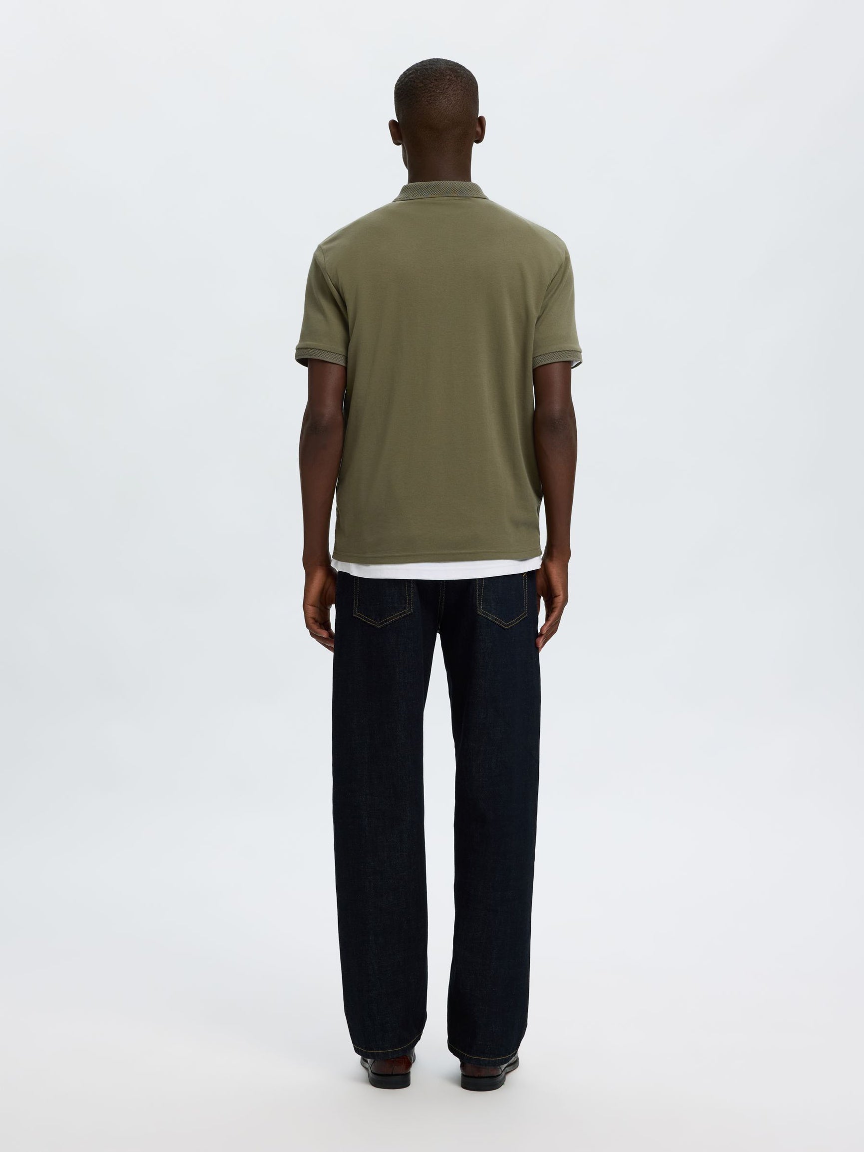 A man viewed from behind wearing an olive green short-sleeve polo shirt and dark blue jeans, standing against a plain white studio background.