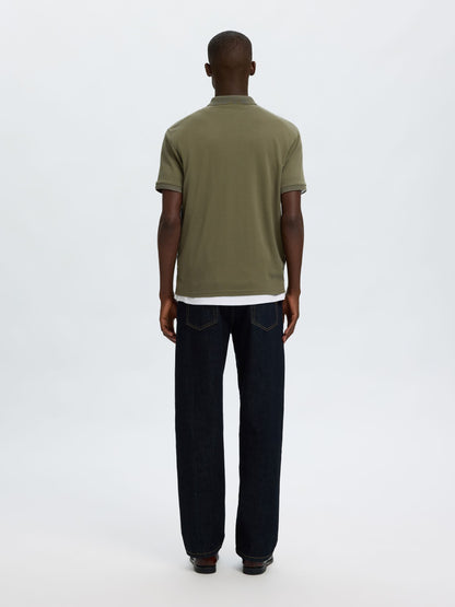 A man viewed from behind wearing an olive green short-sleeve polo shirt and dark blue jeans, standing against a plain white studio background.