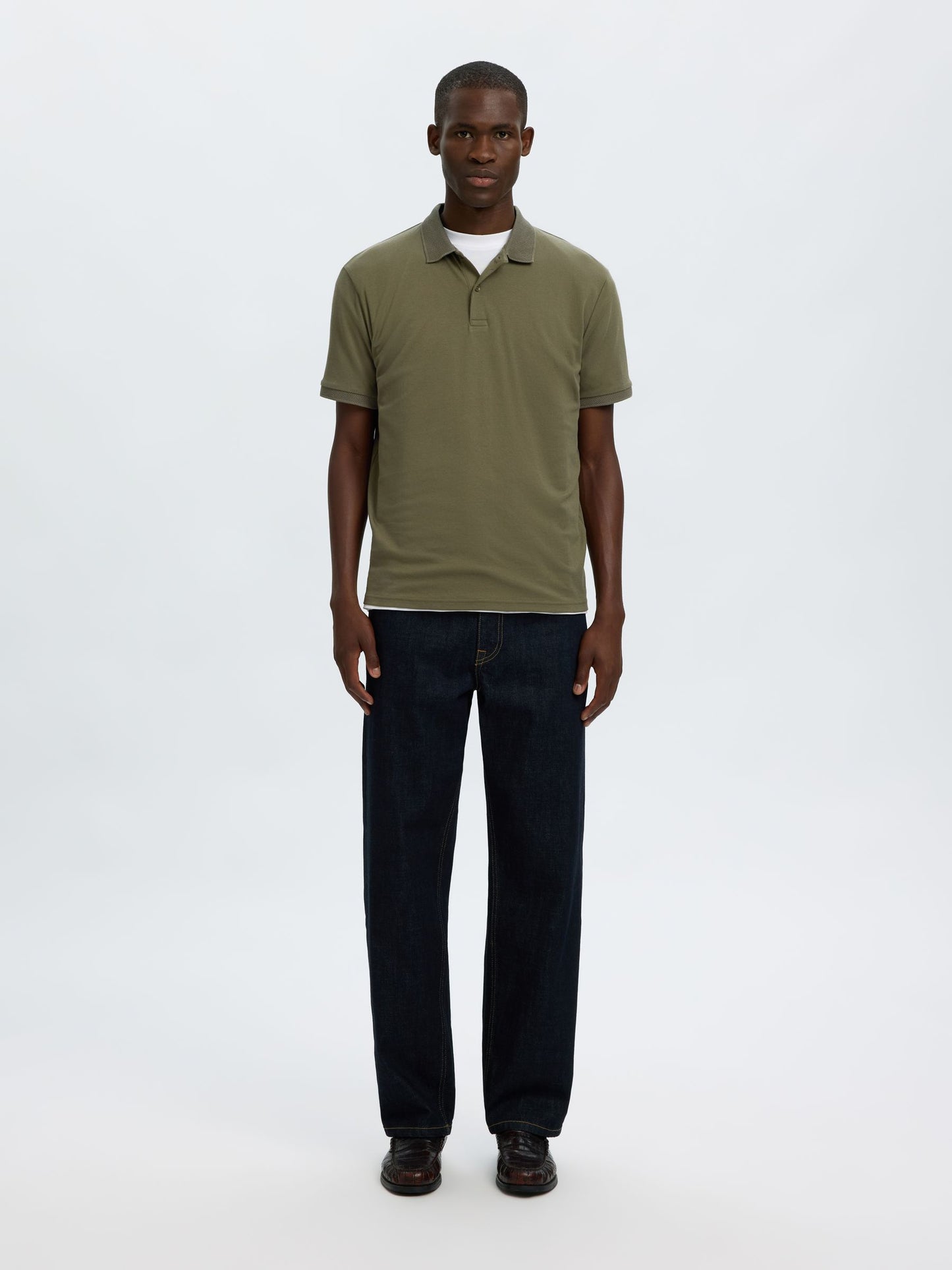 A man modeling a short-sleeve olive green polo shirt with dark blue jeans and brown shoes, standing against a plain white studio background.