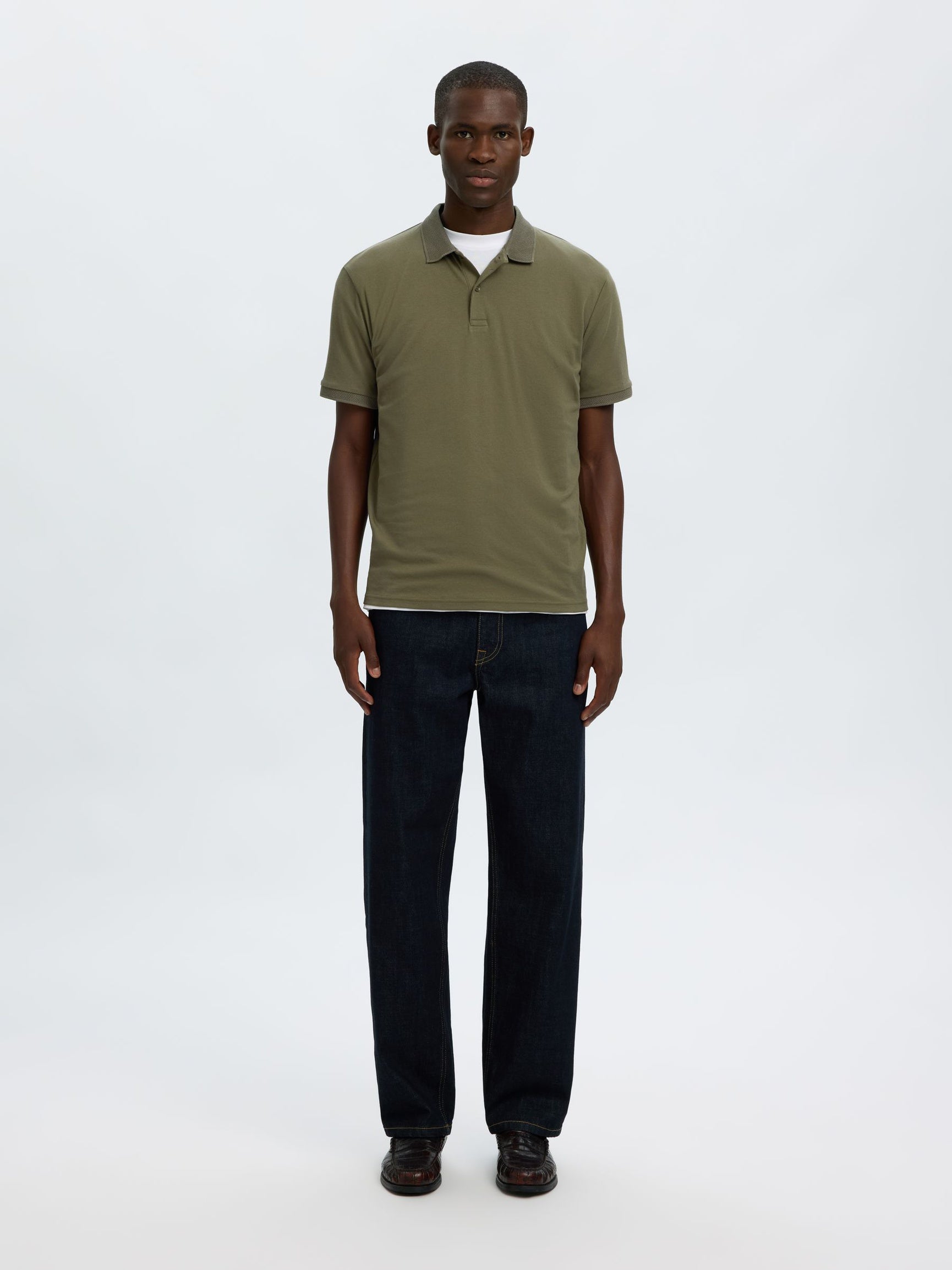 A man modeling a short-sleeve olive green polo shirt with dark blue jeans and brown shoes, standing against a plain white studio background.