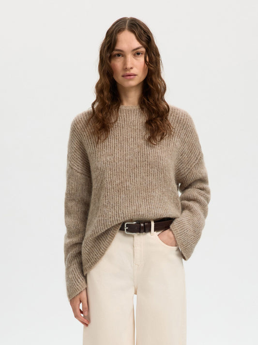 Woman wearing a beige sweater and white pants on a light gray background