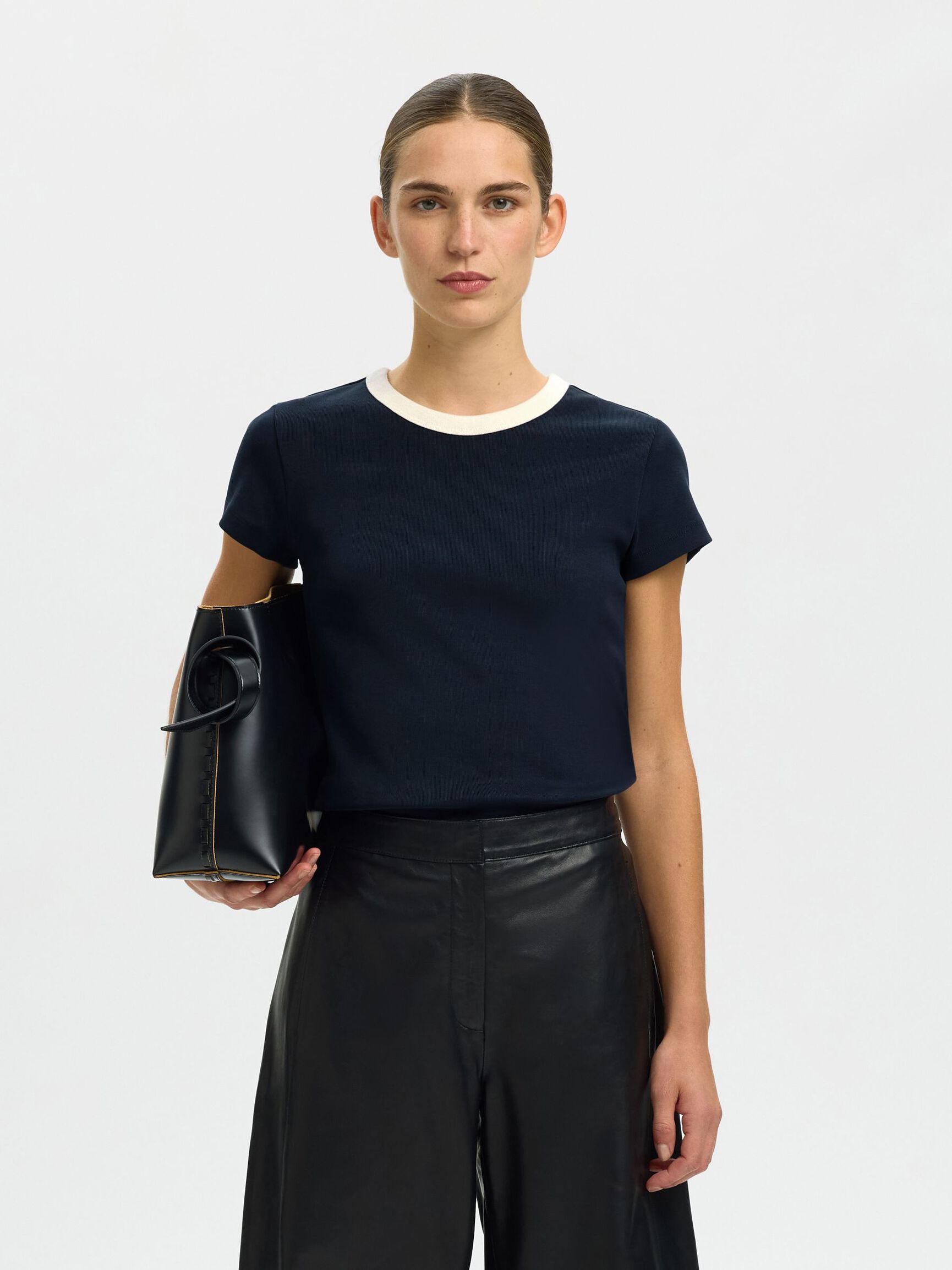 Woman holding a black handbag wearing a navy blue top and black leather pants on a white background