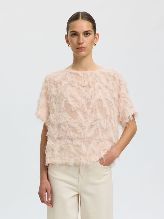 A model wearing a pale pink textured, feather-like short-sleeve top paired with cream-colored trousers, photographed against a seamless light gray studio background.
