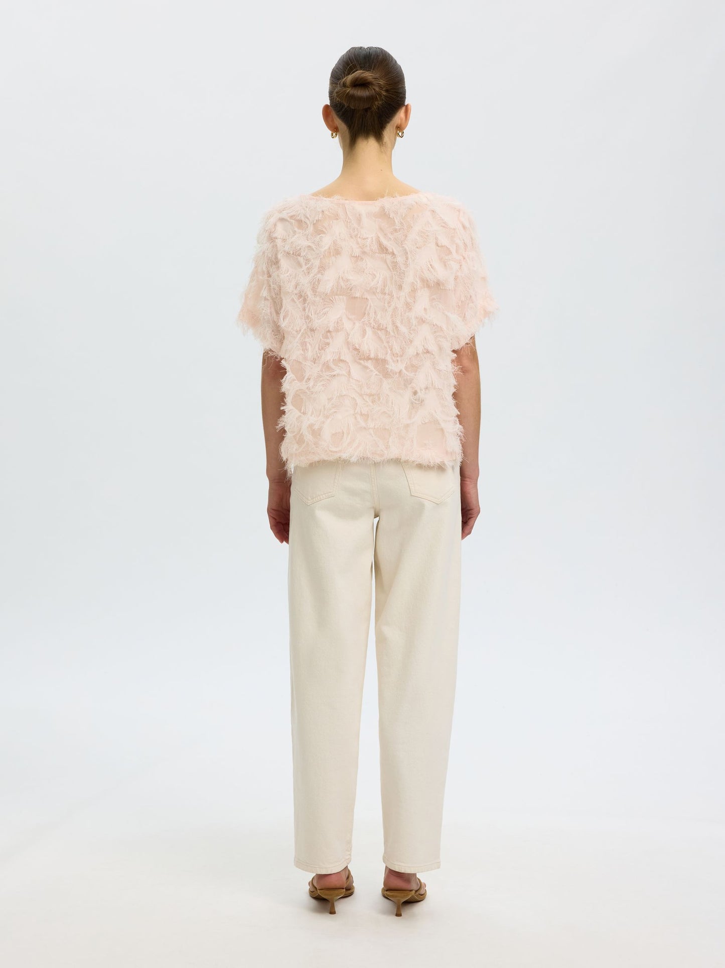 A model shown from the back wearing a light pink feathered ruffled short-sleeve top and cream-colored wide-leg trousers, set against a plain white studio background.