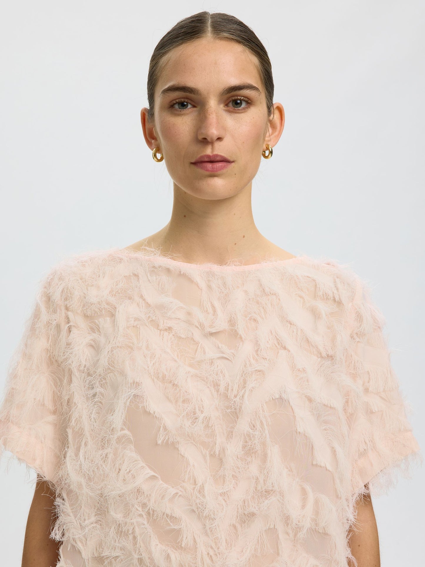 A model wearing a pale pink, short-sleeved sheer mesh top with a textured, feather-like overlay, photographed against a light gray studio background.