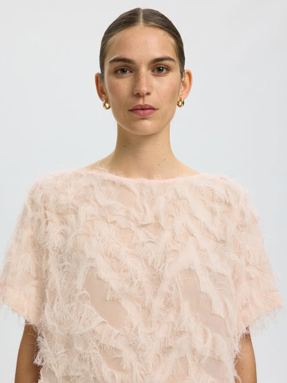 A model wearing a pale pink, short-sleeved sheer mesh top with a textured, feather-like overlay, photographed against a light gray studio background.