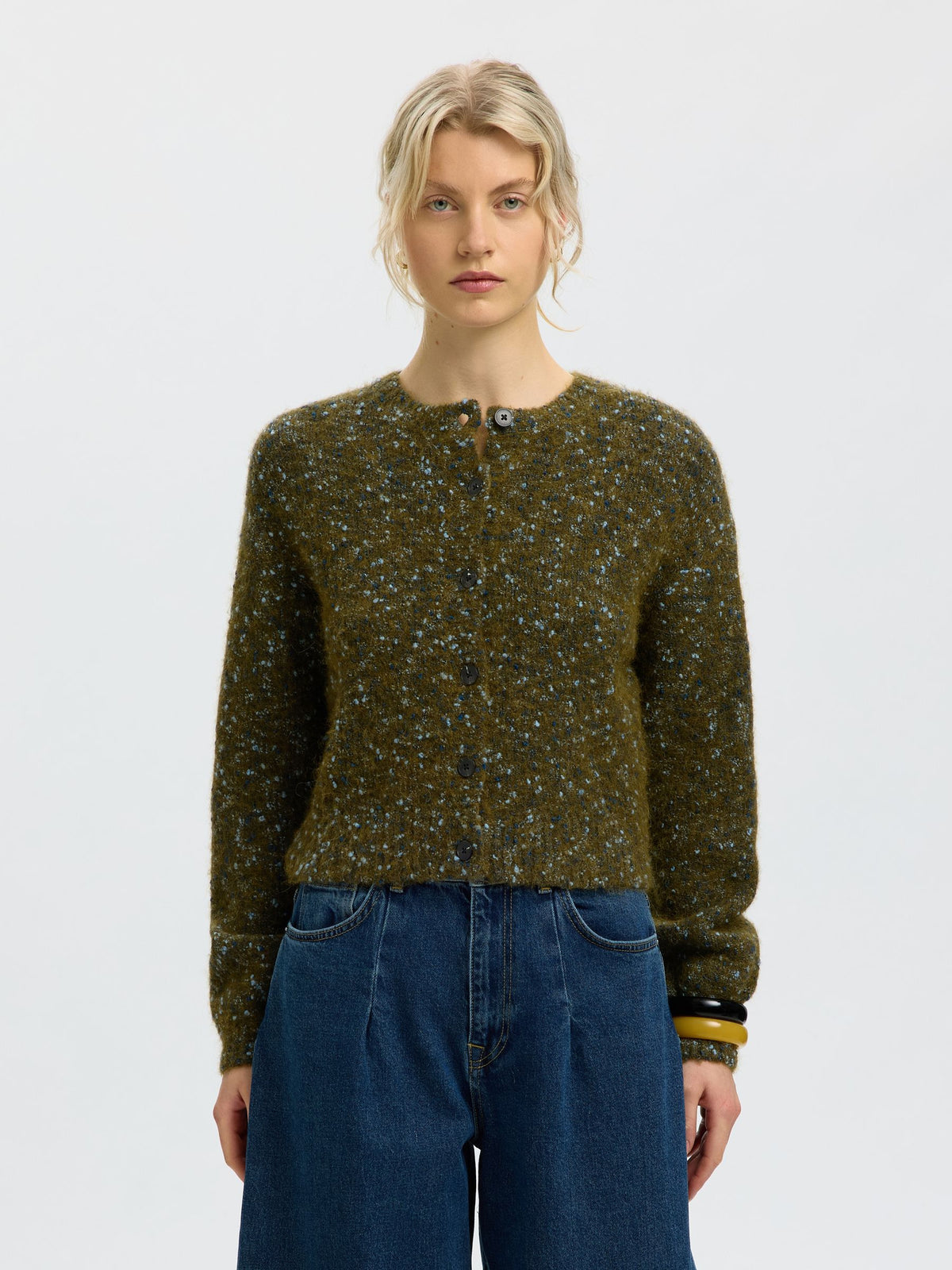 A model wearing a dark olive green knit cardigan sweater with a speckled pattern of blue, white, and yellow flecks, paired with high-waisted blue jeans, against a plain light gray background.
