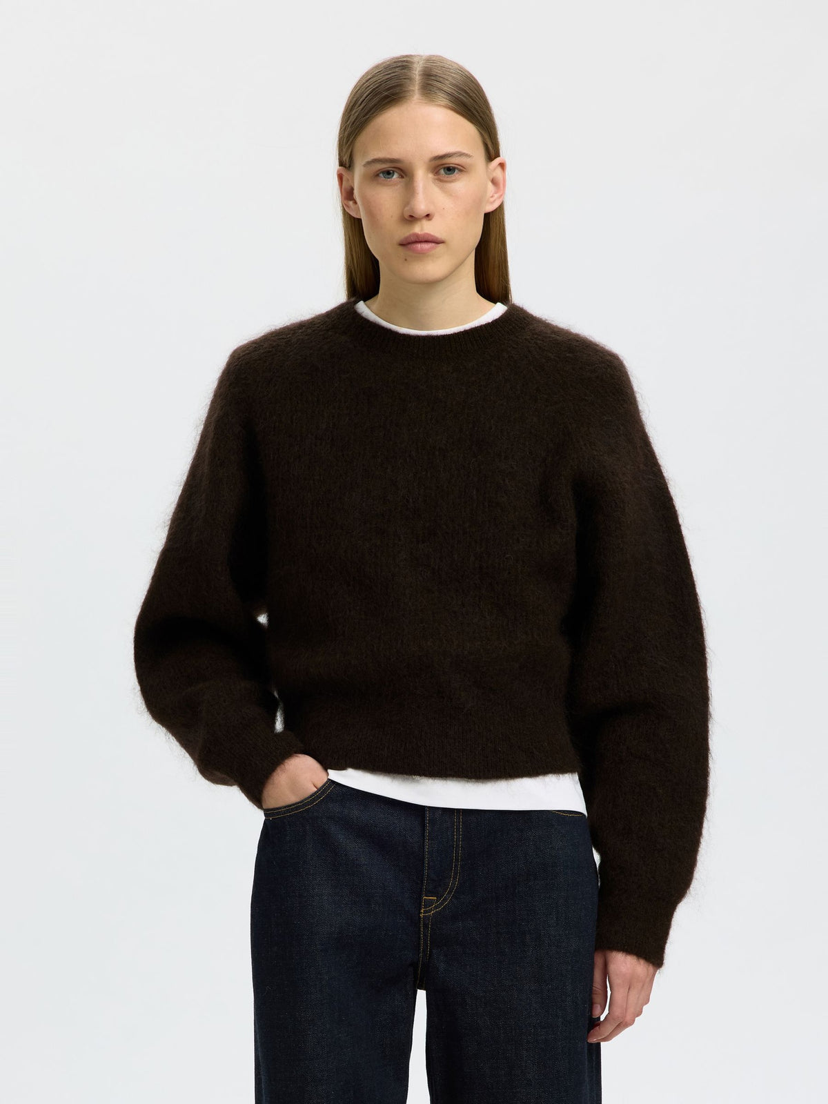 A woman modeling a dark brown fuzzy crewneck sweater over a white undershirt and dark blue jeans, shot against a light gray studio background.