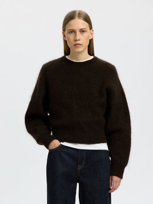 A woman modeling a dark brown fuzzy crewneck sweater over a white undershirt and dark blue jeans, shot against a light gray studio background.