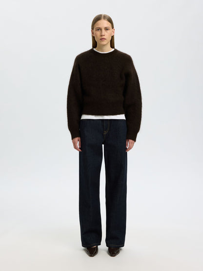 A model stands against a light gray studio background wearing a dark brown knit sweater over a white shirt, dark blue wide-leg jeans, and dark brown shoes.