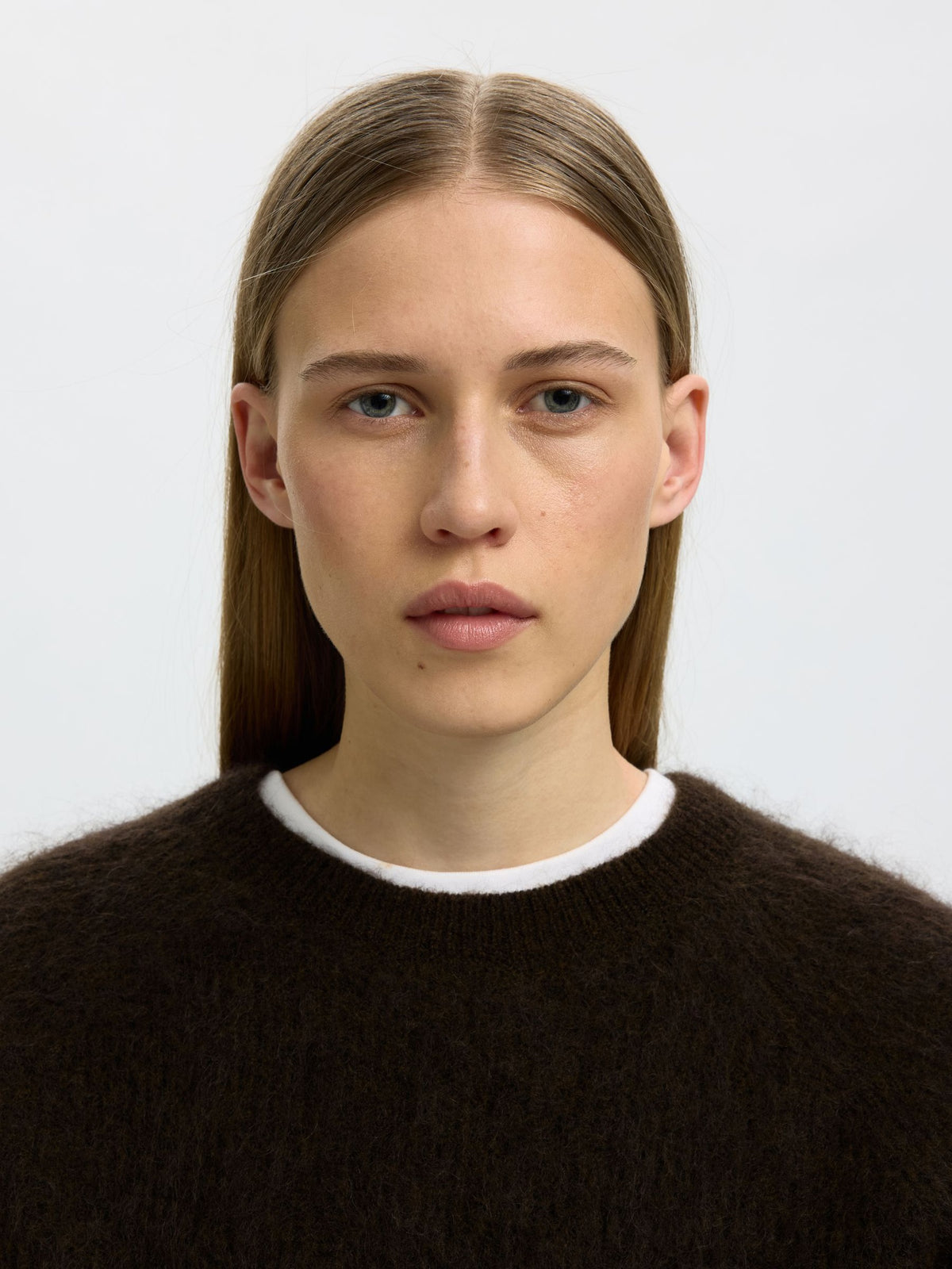 A close-up portrait of a model wearing a dark brown fuzzy knit crewneck sweater with a white ribbed collar, set against a plain light gray background.