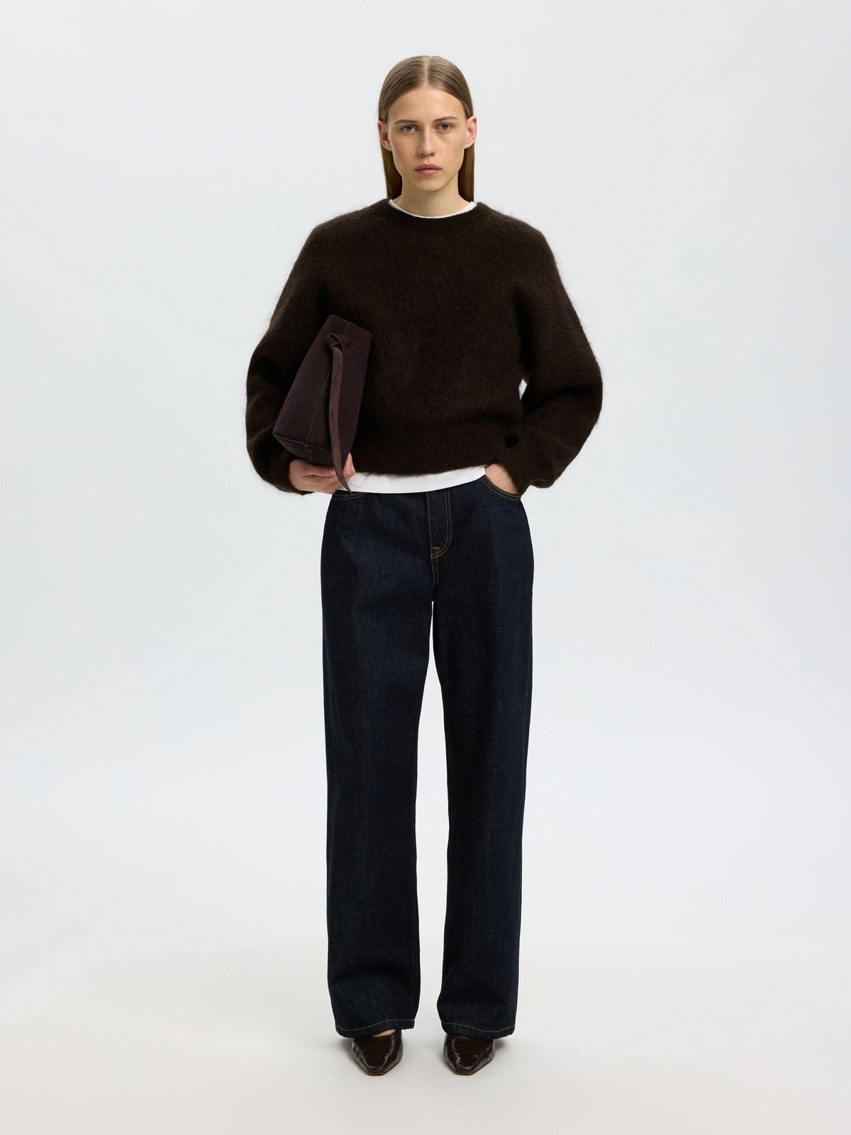 A model stands against a light gray studio background wearing a dark brown fuzzy crewneck sweater over a white top, dark wide-leg jeans, and dark brown shoes, while holding a dark brown envelope-style clutch.