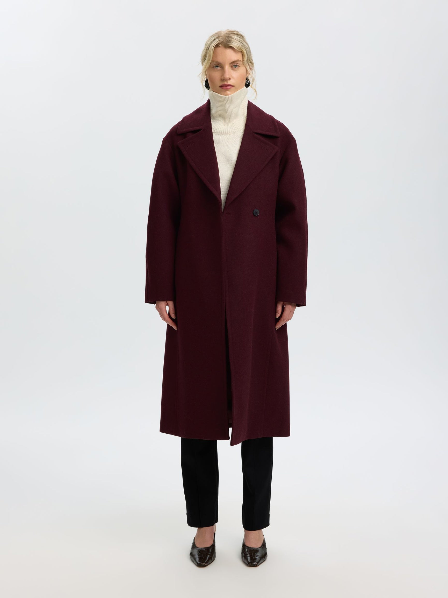 A model stands against a plain white studio background wearing a long burgundy wool-blend coat over a cream turtleneck, black trousers, and dark pointed flats.