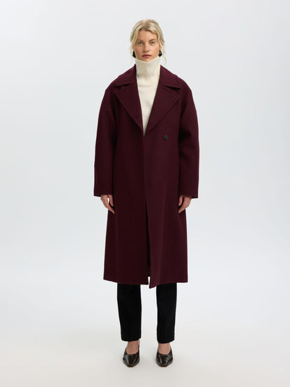A model stands against a plain white studio background wearing a long burgundy wool-blend coat over a cream turtleneck, black trousers, and dark pointed flats.