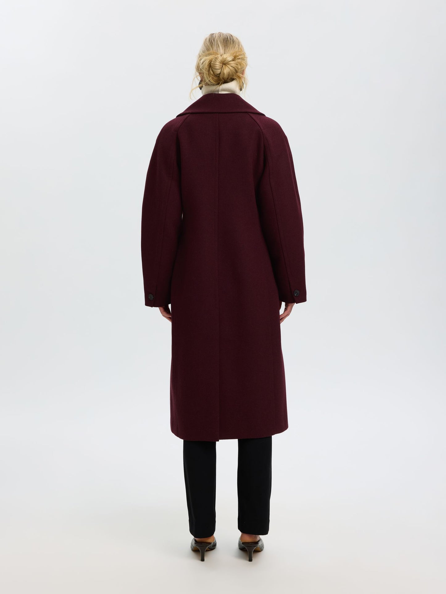 A woman viewed from behind wearing a long burgundy wool coat, black trousers, and brown pointed-toe heels against a plain white studio background.