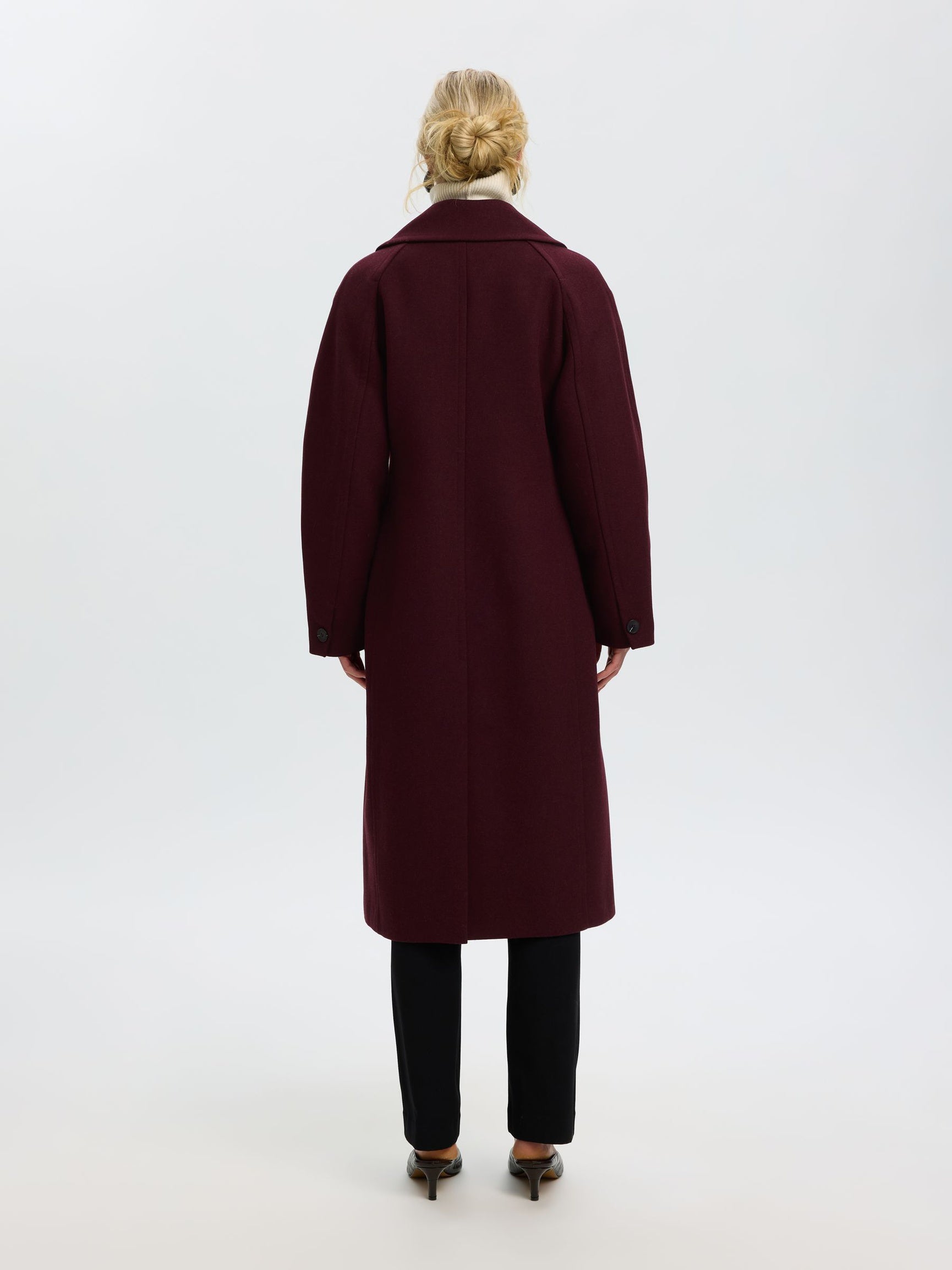 A woman viewed from behind wearing a long burgundy wool coat, black trousers, and brown pointed-toe heels against a plain white studio background.