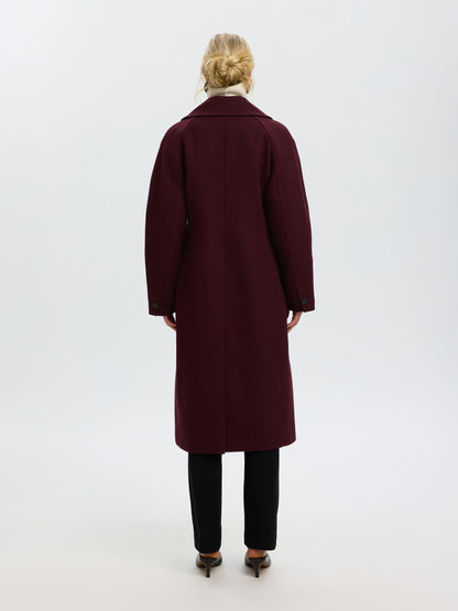 A woman viewed from behind wearing a long burgundy wool coat, black trousers, and brown pointed-toe heels against a plain white studio background.