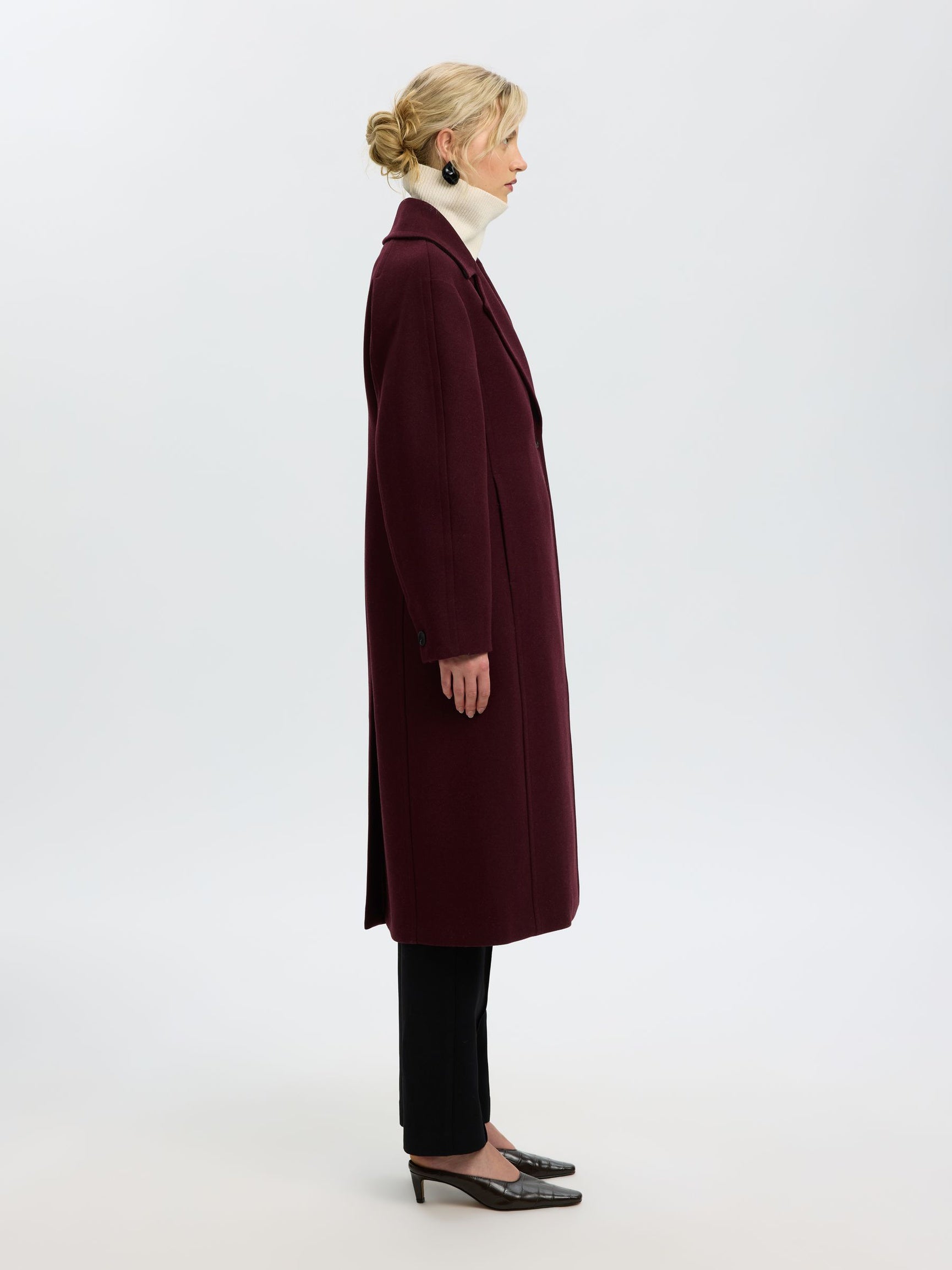 A woman shown in profile wearing a long burgundy coat over a white turtleneck, black pants, and black pointed heels against a plain light gray studio background.