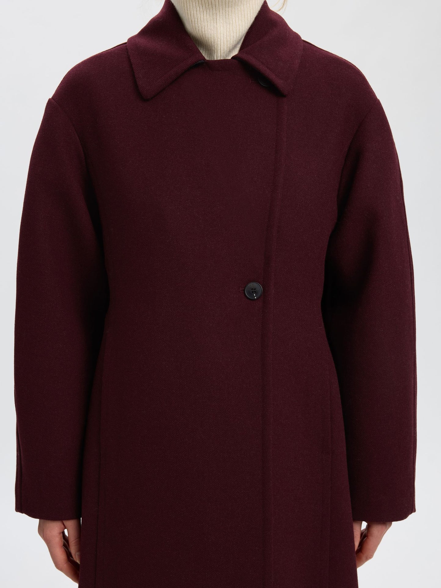 A close-up of a person wearing a dark burgundy wool coat with a single button closure, layered over a cream ribbed turtleneck, against a light gray background.
