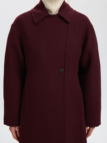 A close-up of a person wearing a dark burgundy wool coat with a single button closure, layered over a cream ribbed turtleneck, against a light gray background.