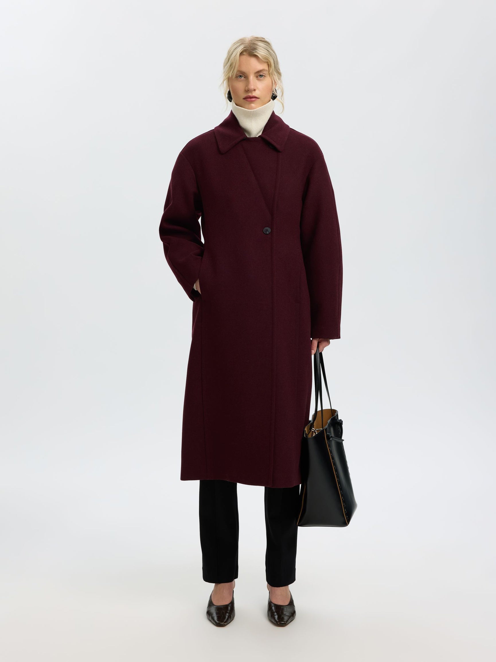 A woman stands against a light gray studio background wearing a long burgundy overcoat over a white turtleneck, black pants, and black shoes, while holding a black tote bag.