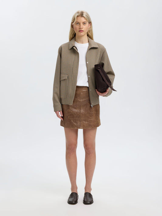A model stands against a light gray studio background wearing a brown and beige checkered jacket over a white top, a brown mini skirt, and black shoes, while holding a dark brown clutch.