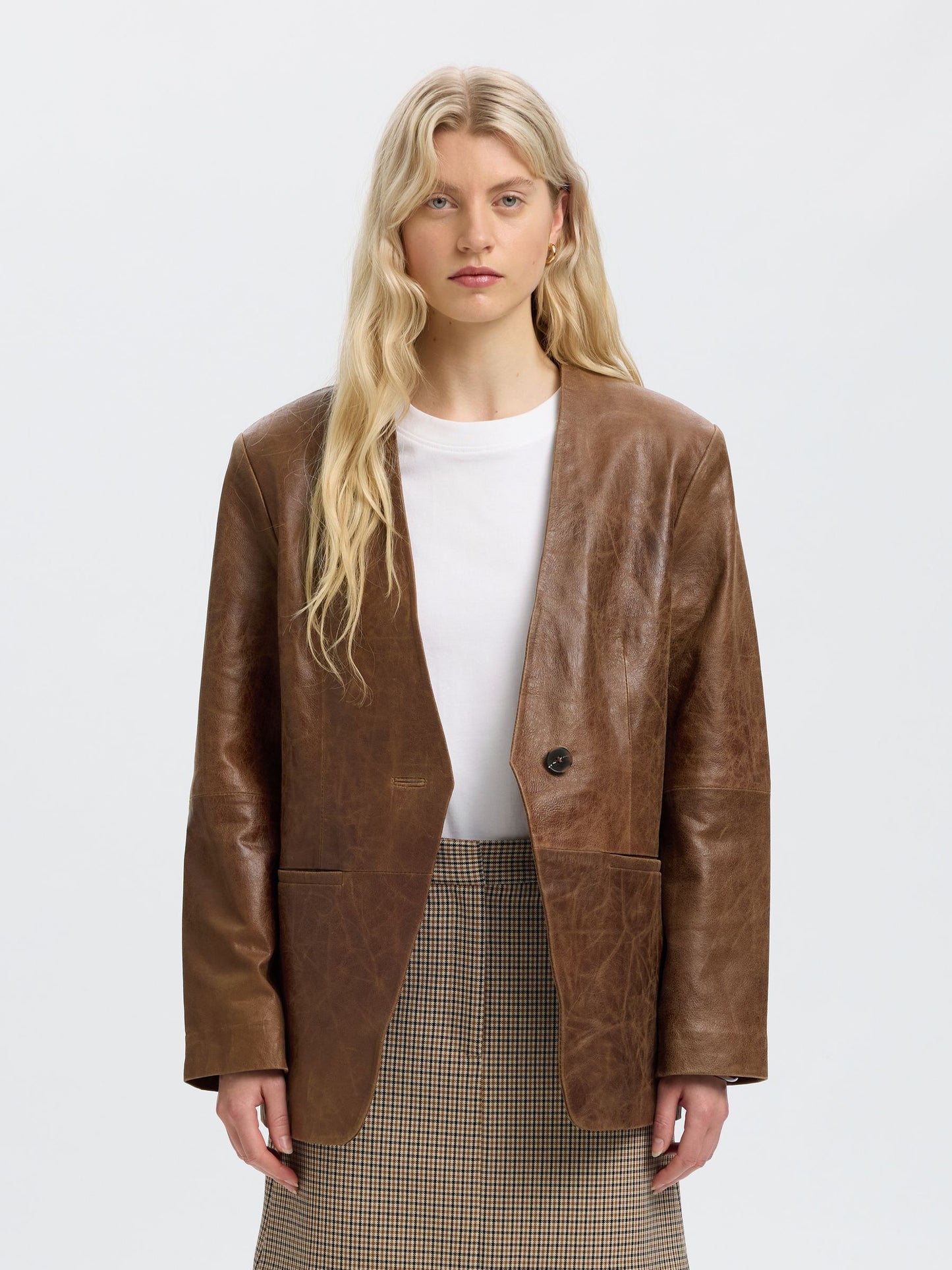 A blonde model wearing a brown leather blazer over a white top and a brown-and-beige plaid skirt, photographed against a light gray studio background.
