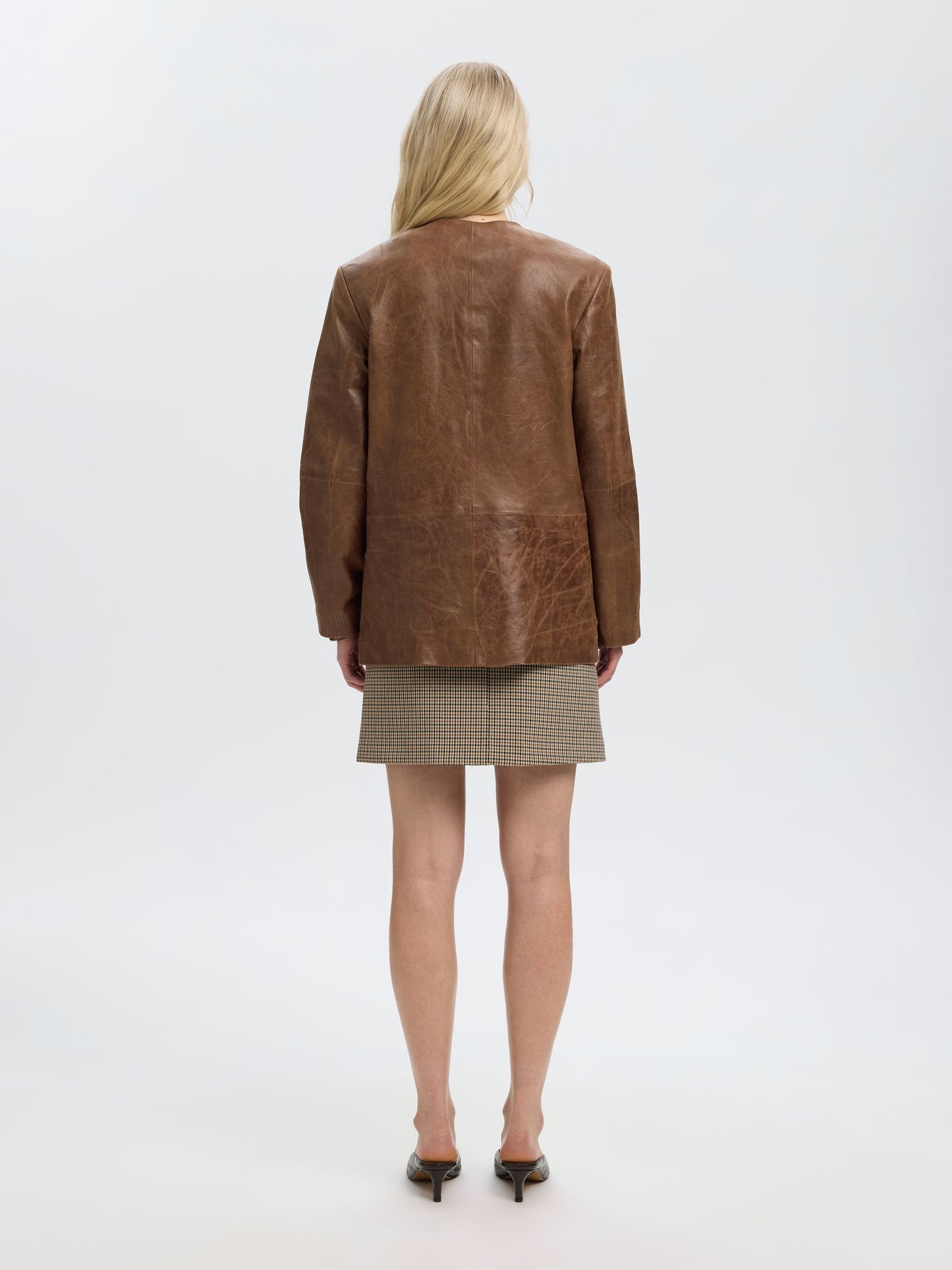 A woman shown from the back wearing an oversized brown leather jacket over a beige and black checkered mini skirt, paired with black pointed-toe heels, against a plain white studio background.