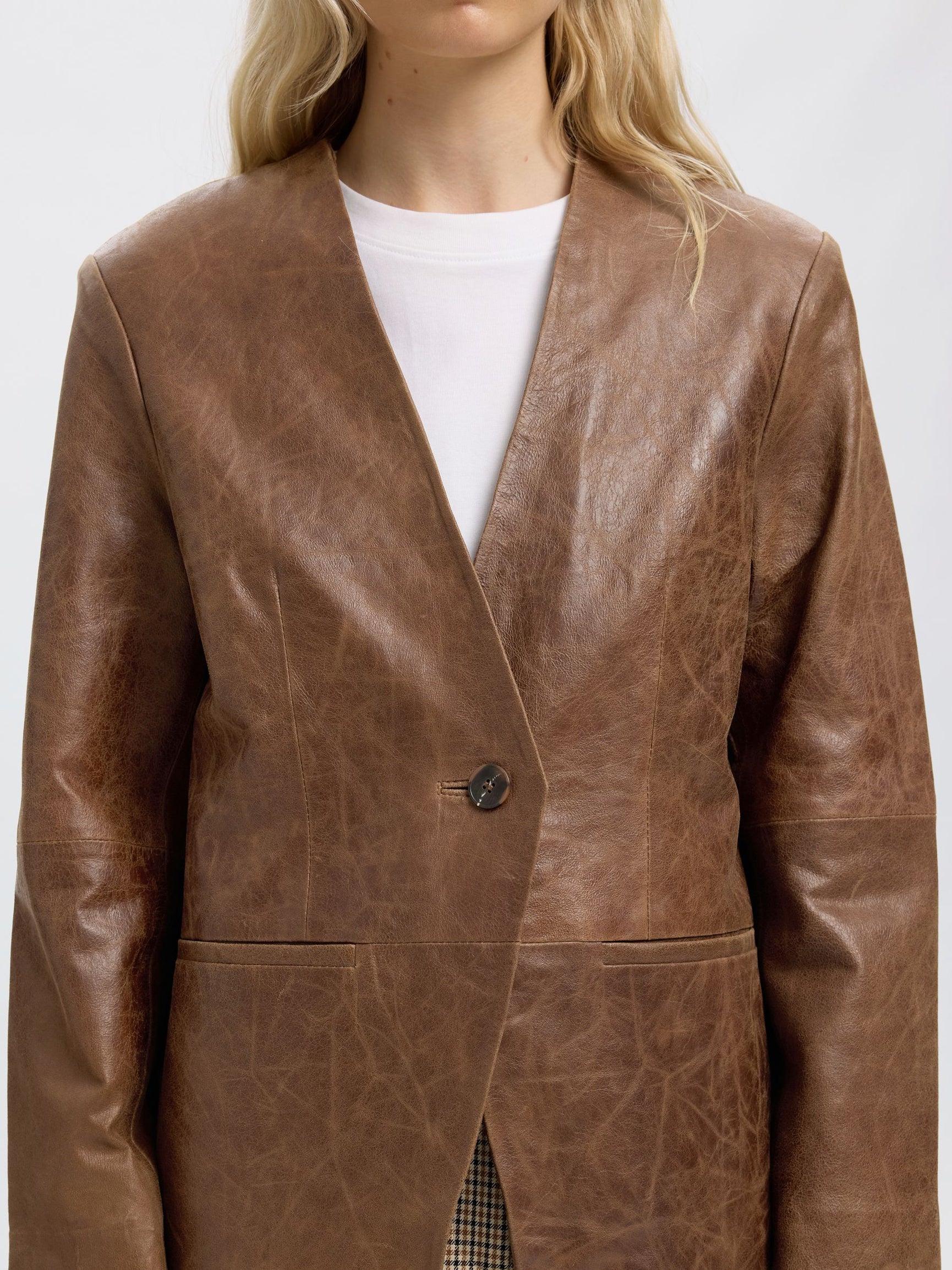 A close-up of a model wearing a brown textured faux-leather blazer over a white crewneck t-shirt, against a plain light gray background.