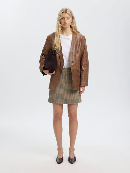 A blonde model stands against a light gray studio background wearing a brown mixed-media jacket with suede-like panels, a white top, a brown and beige houndstooth mini skirt, and dark pointed flats. She holds a dark brown clutch.