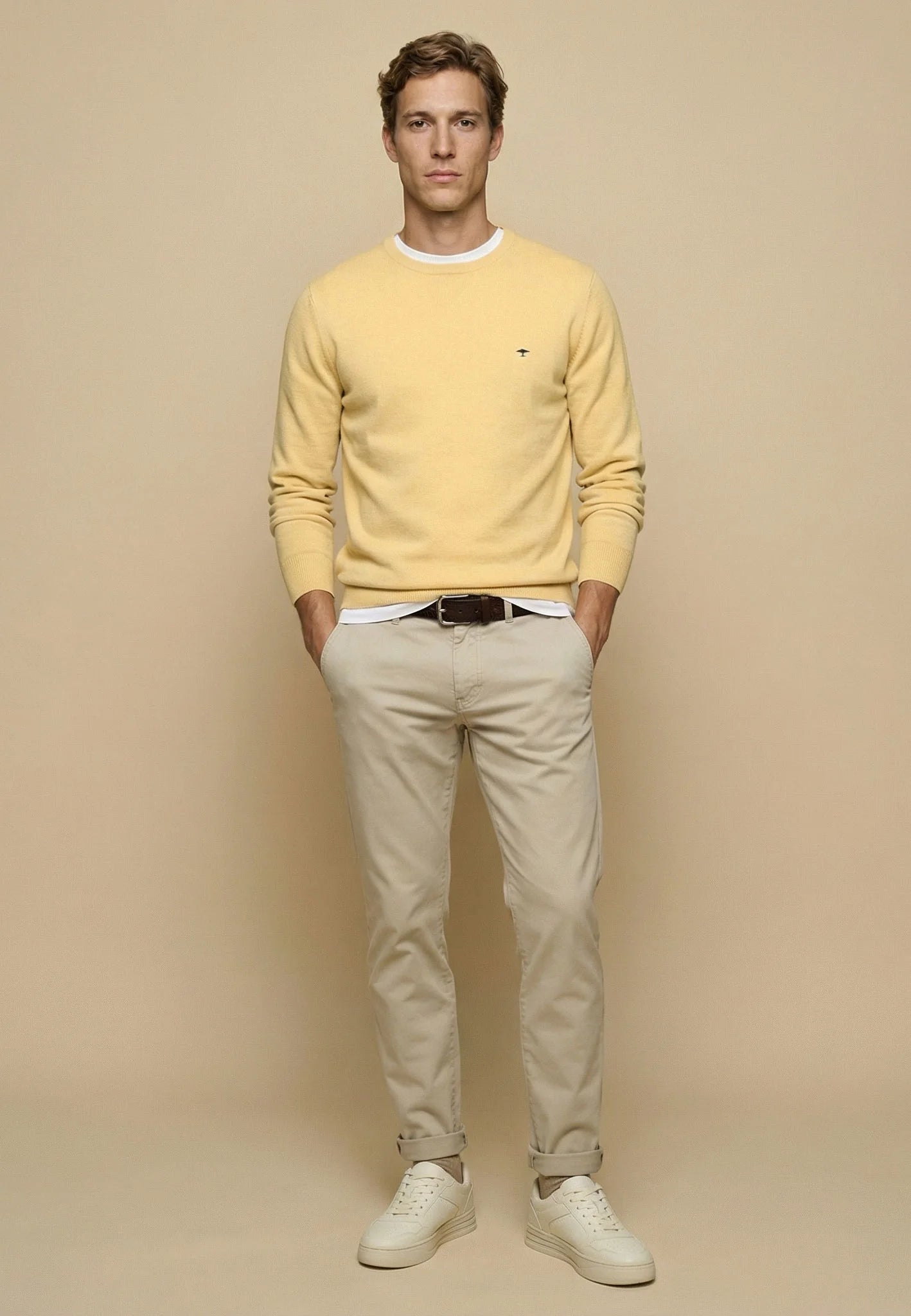 A man modeling a pale yellow crewneck sweater, beige trousers, and white sneakers against a solid beige studio background.