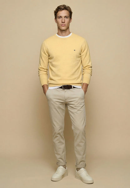 A man modeling a pale yellow crewneck sweater, beige trousers, and white sneakers against a solid beige studio background.