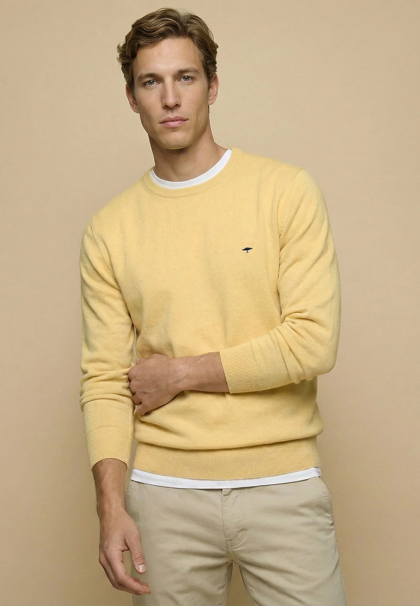 A man wearing a pale yellow crewneck sweater with a white undershirt and khaki pants, standing against a solid beige background.